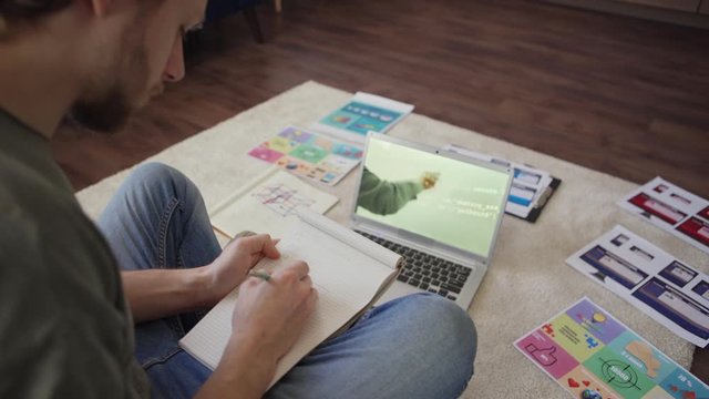 Tilt up and down panning shot of male college student studying programming online listening to video conference with teacher sitting on floor at home