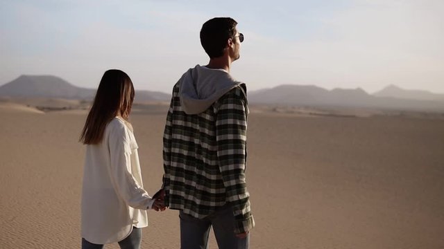 Happy couple holding hands walking in romantic relationship under sun and blue sky in desert. Two young lovers walking by desert sand in casual clothes and smiling