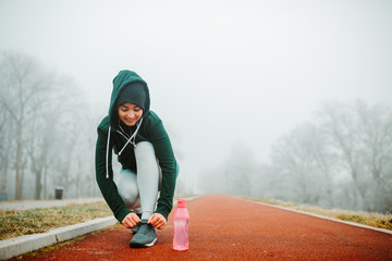 Close photo of young fit woman tying shoe laces. Pink water bottle is on the ground next to her.