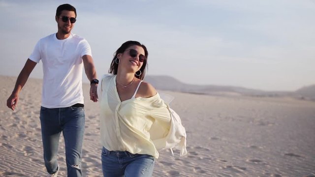 Couple holding hands running having fun under sun in playful and romantic relationship under sun and blue sky in desert. Two young lovers cheerful together on romance in summer. Wearing casual clothes