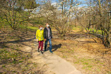Obraz premium Man and woman couple in protective medical masks walk through the park on a sunny spring afternoon.