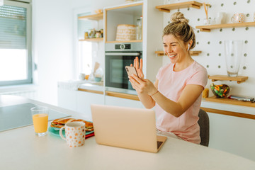 Young woman is having video call with her friends while sitting in kitchen at home. Blonde girl is using wireless headphones and smartphone to have video call with friends.