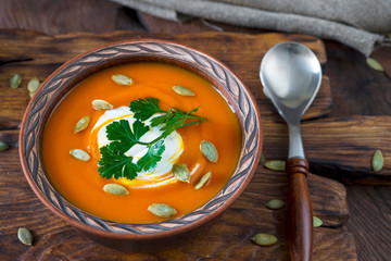 Pumpkin soup in a clay bowl, spoon, whole pumpkins and burlap on a dark wooden background.