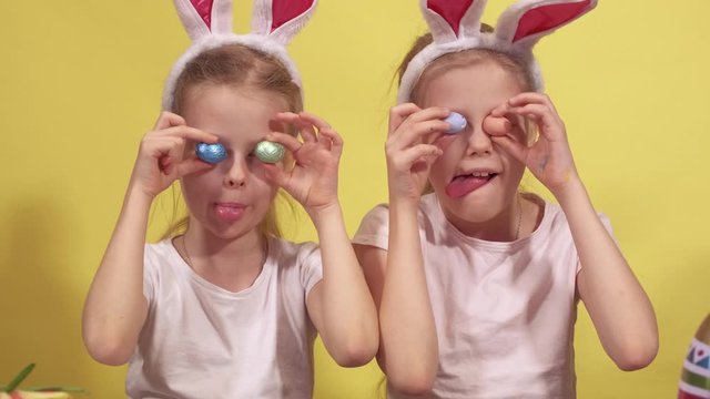 Cute sisters with bunny ears keeping colorful quail eggs near eyes and showing tongues while preparing for Easter celebration against yellow background