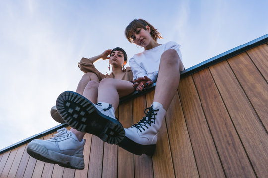 Two Girlfriends Sitting On a Wooden Wall Looking At Camera.