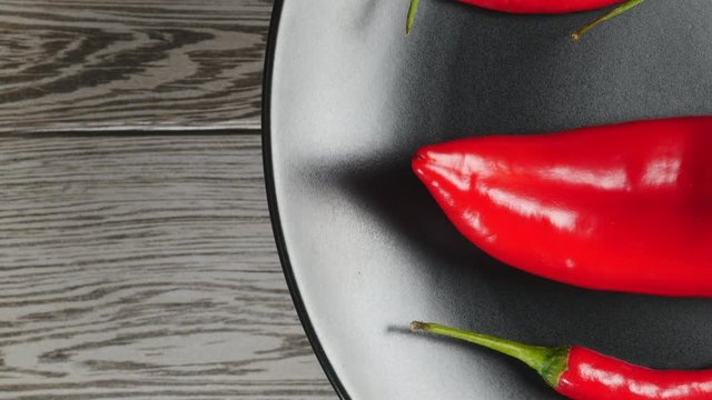 Top View Of Fresh Raw Red Chili Peppers And Ramiro Pepper Rotating On A Black Plate On A Gray Wooden Background On The Right Side.