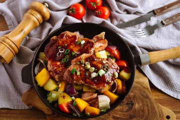 Black pan with fried slices of pork meat, with chopped vegetables: pumpkin, bell pepper, green onion. On a wooden table, a board, a salt mill, tomatoes, a fork and a knife, a gray cloth napkin.