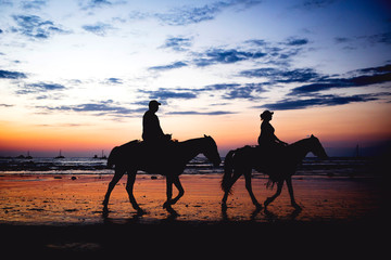 Silhouette of two people riding horses on the beach at sunset