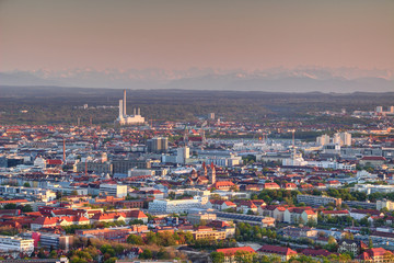 Aerial view of modern European city outskirts in evening light with industrial, commercial and residential buildings, power plant with chimneys and Bavarian Alps in background, Munchen Bayern Germany