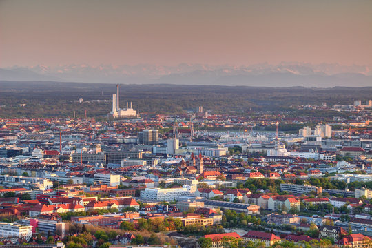 Aerial view of modern European city outskirts in evening light with industrial, commercial and residential buildings, power plant with chimneys and Bavarian Alps in background, Munchen Bayern Germany