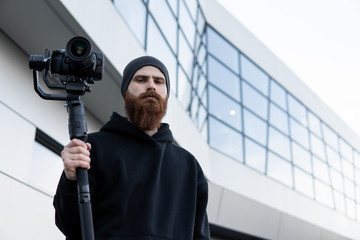 Bearded Professional videographer in black hoodie holding professional camera on 3-axis gimbal stabilizer. Filmmaker making a great video with a professional cinema camera. Cinematographer.