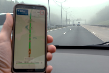 Selective focus. Hand holds a mobile phone with a navigation application in a car. View of the highway in fog and rainy weather. Low visibility while driving. Glonass navigator