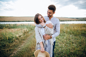 Happy couple in love hugging, kissing and smiling against the sky in field. Hat in girl's hand