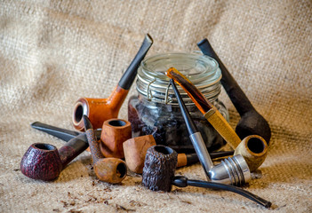 Pile of Unique old pipes with a jar of loose tobacco on a burlap background 