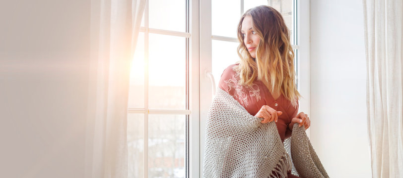 Thoughtful blonde european female wearing beige dress sits on a windowsill and looks outside