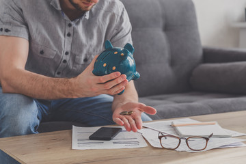 Man sitting at a table full of unpaid bills shakes out the last penny from the piggy bank. Spend your last savings. Unemployment, poverty, bankruptcy concept.