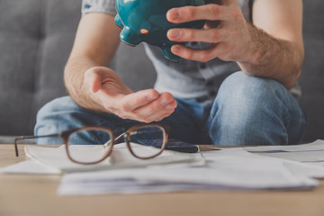 Man sitting at a table full of unpaid bills shakes out the last penny from the piggy bank. Spend...