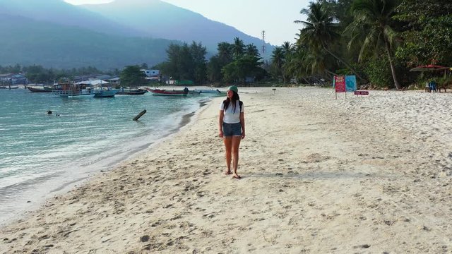 Trendy girl walking slowly on white sand watching calm sea water washing tranquil exotic beach on Ko Pha Ngan island in Thailand