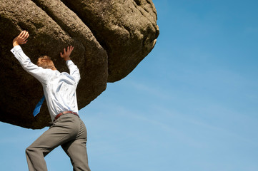 Strong businessman struggling to lift massive boulder into blue sky copy space