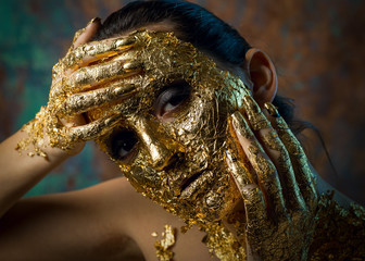 Girl with a mask on her face made of gold leaf. Gloomy studio portrait of a brunette on an abstract background.