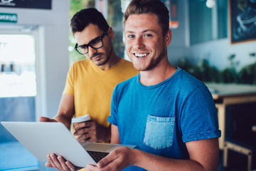 Portrait of cheerful prosperous male freelancer looking at camera holding laptop computer while his colleague watching online video on browsed web page spending time in coworking space together.