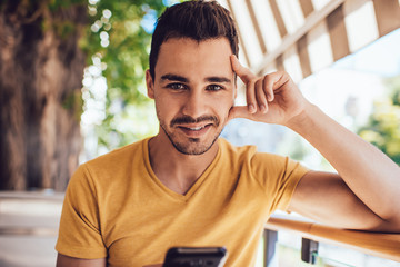 Pleased young man in casual clothing chatting on smartphone at outdoors cafe