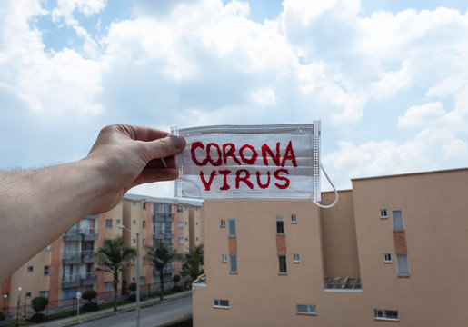 Closeup to a Facemask with red "CORONAVIRUS" inscription holding by a male hand with neighborhood and residential buildings in sunny day at background.  