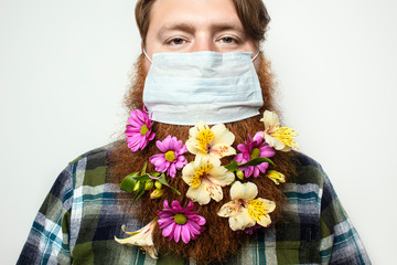 A man with a red hair and a red beard wearing a face mask.  A beard decorated with flowers for spring holidays at quarantine. A bearded man with a face mask protecting himself from COVID-19 epidemic.