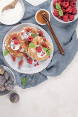 Pancakes with raspberries, figs, yogurt, coconut zest, honey and mint leaves on a plate.
