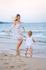 A blonde mother in a white dress plays with her daughter in the waves near the blue sea and laughs.