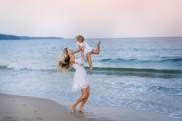 Fototapeta na wymiar A blonde mother in a white dress throws her daughter in her arms by the sea and laughs.
