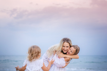 Fototapeta na wymiar Mom with daughters blondes in white dresses laugh and hug near the blue sea on the beach at sunset.
