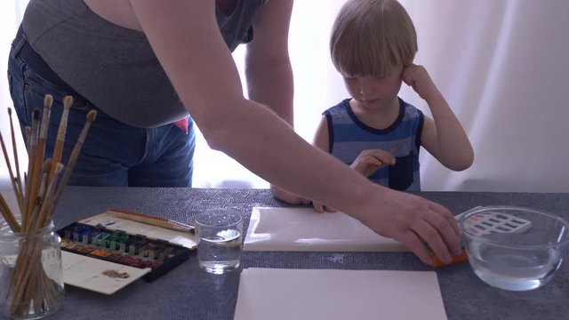 adult father teaches to paint lovely blond child with watercolors. Paints in cuvettes, brushes, water for wetting paper. boy likes to spend time with his father. Wet paper produces beautiful patterns