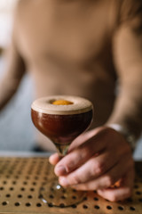 Bartender holding on his hand glass with espresso martini cocktail decorated with slice of orange zest. Smooth image with shallow depth of field.