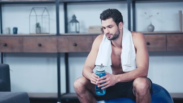 tired sportsman with towel on neck sitting on fitness ball and drinking water