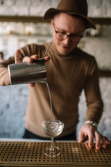 Bartender in a hat pouring a cocktail from a mixing pitcher into a cooled glass. Smooth image with shallow depth of field.