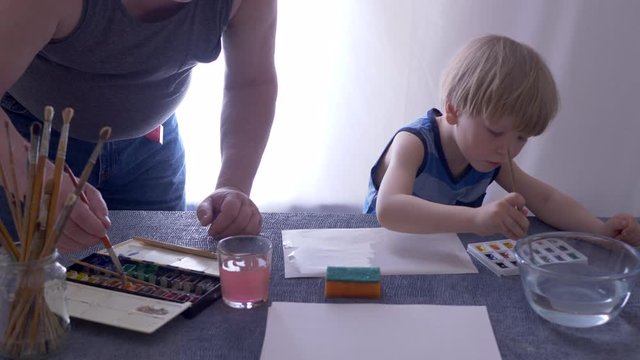 adult father teaches to paint lovely blond child with watercolors. Paints in cuvettes, brushes, water for wetting paper. boy likes to spend time with his father. Wet paper produces beautiful patterns