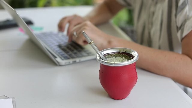 Woman working on laptop and drinking yerba mate tea in home office. Female hands typing on laptop, drinking hot yerba mate tea. Slow Motion.