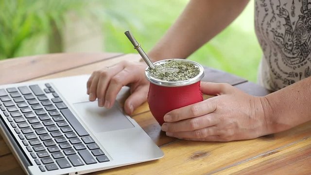 Woman working on laptop and drinking yerba mate tea in home office. Female hands typing on laptop, drinking hot yerba mate tea. Slow Motion.