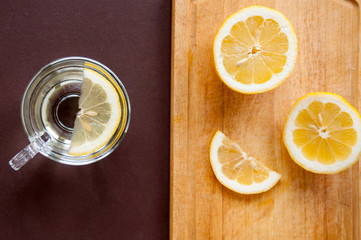 On a wooden cutting Board, slices of lemon and a transparent Cup with lemon on a brown background, top view