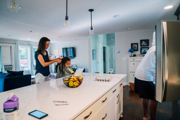 Girl studying on laptop while mom fixes her hair at kitchen counter, dad getting food out of fridge in background