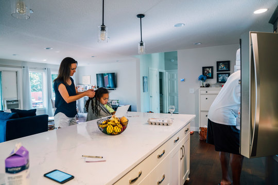 Girl studying on laptop while mom fixes her hair at kitchen counter, dad getting food out of fridge in background