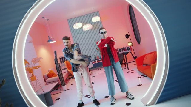 Zoom in shot of two professional male dancers in eccentric outfits performing vogue in vintage studio with pink lighting and illuminated round doorway
