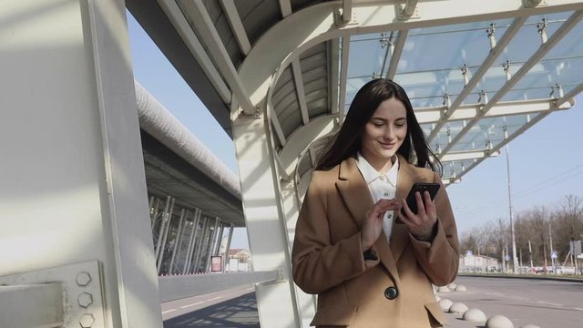A youngSmiling curly woman wearing trendy sunglasses walks down the central city street and uses her phone. Pretty summer woman in white jacket walks down the str lady using a mobile phone outdoor. 
