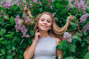 Portrait of a charming blond woman wearing beautiful white dress standing next to lilac bush with hair on the leaves.