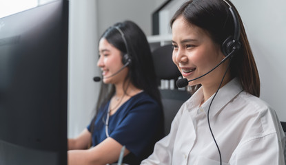 Smiling Asian business woman and team working in call center at office desk with headset and computer.