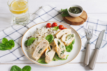 Chicken fillet roll with spinach. Garnished with long-grain wild rice. Cherry tomatoes, pesto sauce, a glass of water and a slice of lemon. Checkered cloth, knife and fork. On a white wooden table.