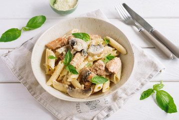 Penne with slices of chicken and turkey and mushrooms in a creamy sauce, grated cheese and fresh basil leaves. On a white wooden table and a tissue napkin. Knife and fork.