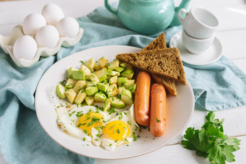 Fried eggs for breakfast on a white plate. Fried eggs with sausages and zucchini, two slices of rye bread, parsley. White wooden background and blue fabric napkin.