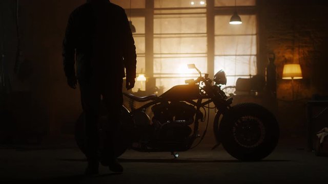 Custom Bobber Motorbike Standing in an Authentic Creative Workshop. Rider Gets on a Bike and Starts It. Vintage Style Motorcycle Under Warm Lamp Light in a Garage.